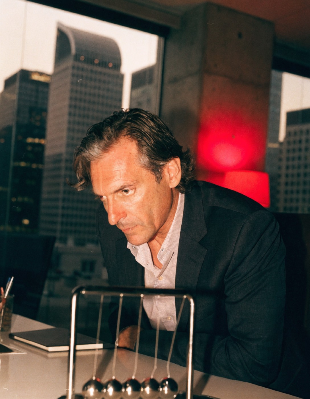 An executive watching a Newton's cradle on his desk, red lamp behind him, waiting on a number that should already be here